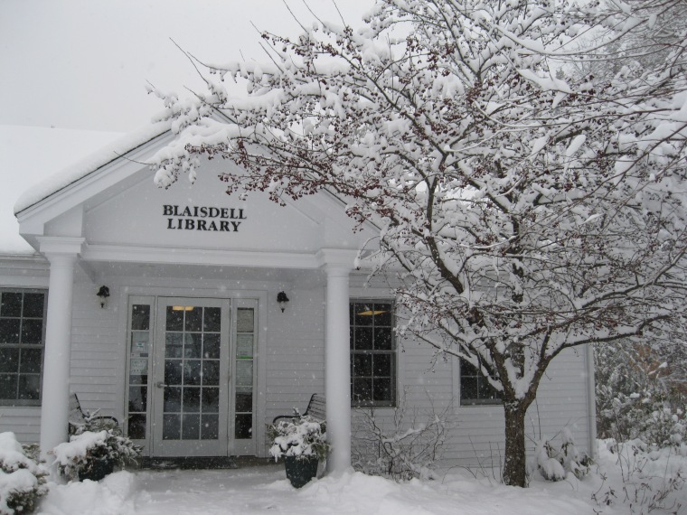 A photo of the exterior of the Blaisdell Memorial Library. It is snowing, and a later of fluffy snow already covers the tree and the ground.